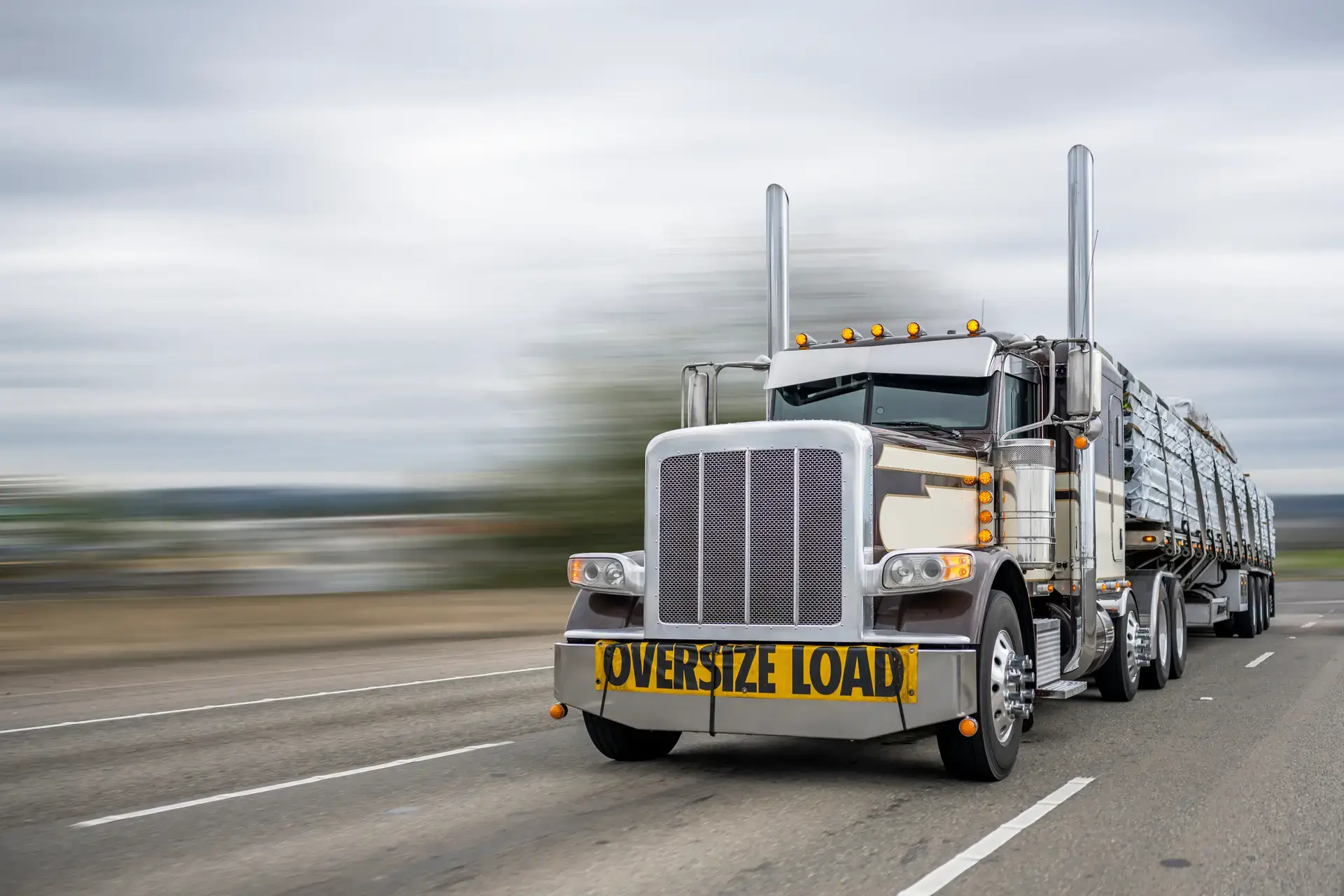 Classic dark long haul big rig bonnet semi truck tractor with oversize load sign on the front transporting fixed with slings lumber on the flat bed semi trailer running on the wide highway road